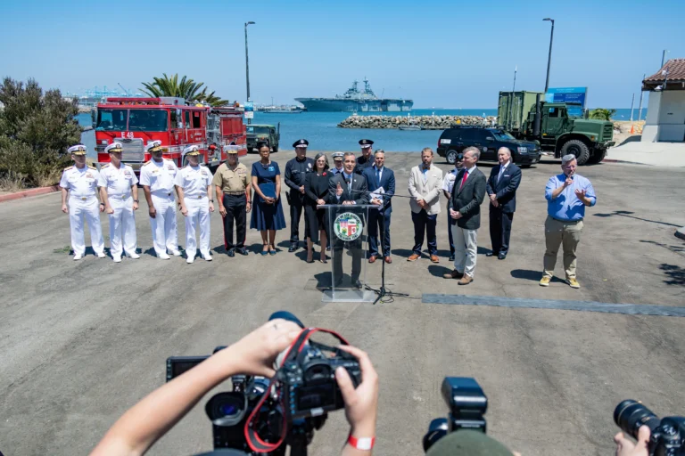 A group of officials and military personnel stand at a podium near the waterfront, with fire trucks, military vehicles, and a naval ship in the background. Photographers capture the scene from the foreground. A group of officials and military personnel stand at a podium near the waterfront, with fire trucks, military vehicles, and a naval ship in the background. Photographers capture the scene from the foreground.