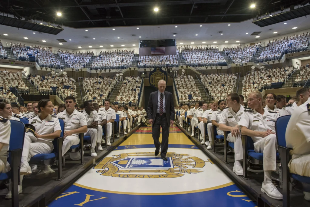 A man in a suit walks down an aisle in a large auditorium filled with people in white uniforms, with a navy emblem on the floor and many seated attendees in the background.