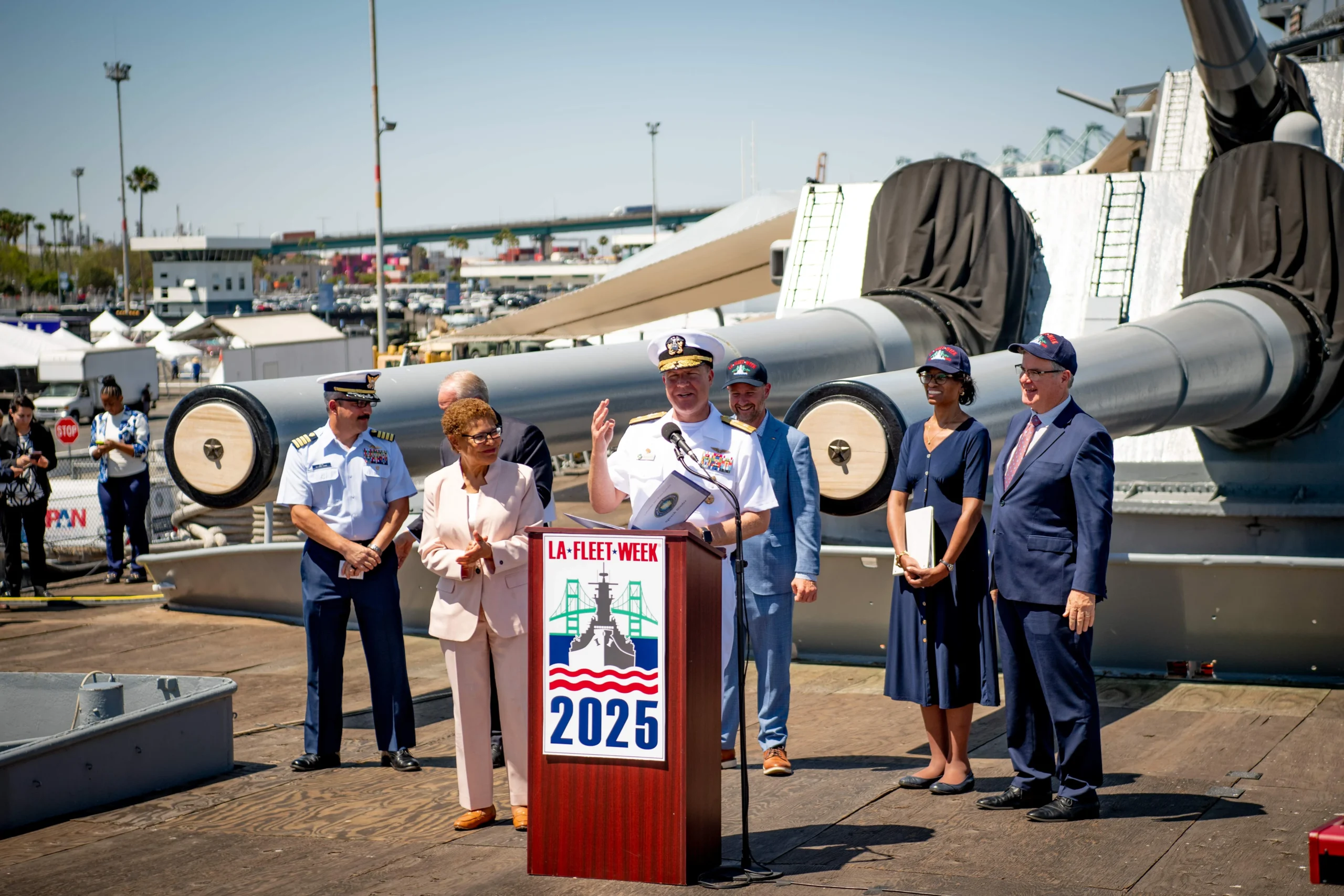 A naval officer speaks at a podium labeled LA Fleet Week 2025, surrounded by military personnel and officials on the deck of a battleship, with large cannons and a port in the background—a powerful scene honoring Navy Legacy and the dedicated commitment to Preserving the Watch.