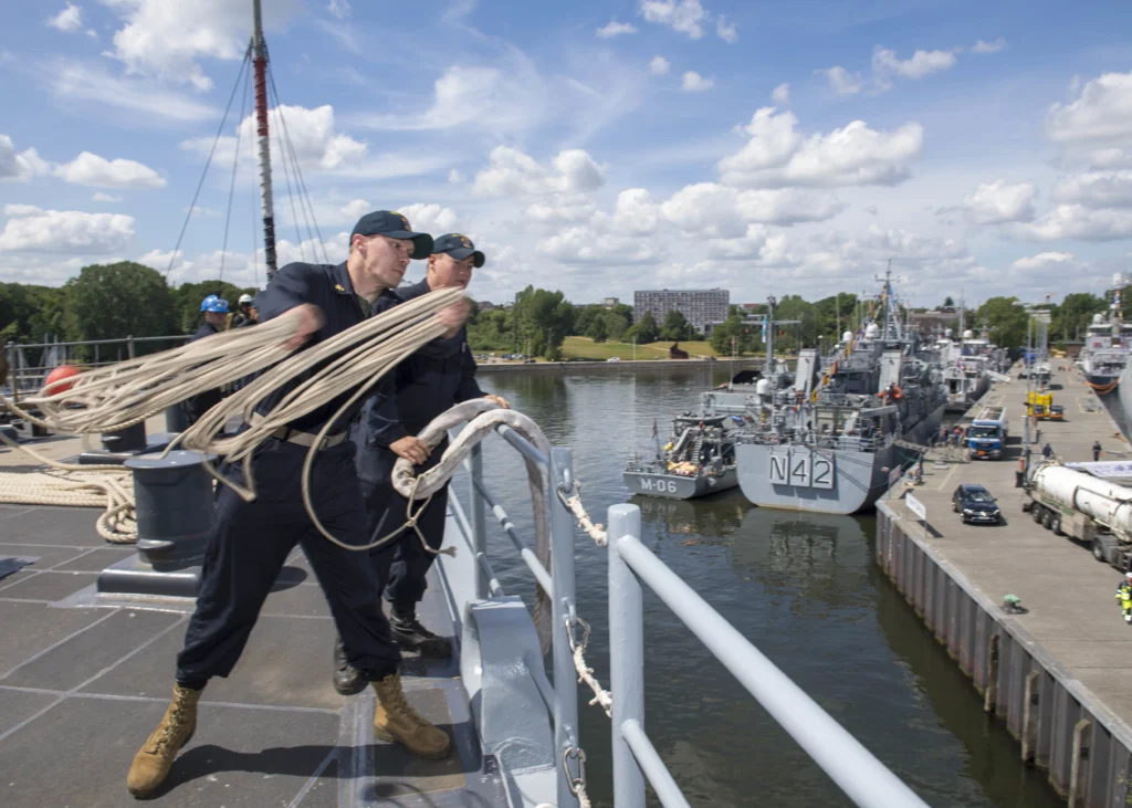 Two sailors in uniform stand on a ship; one throws a rope toward the dock where naval vessels are moored. The scene takes place on a sunny day with partly cloudy skies and buildings visible in the background.