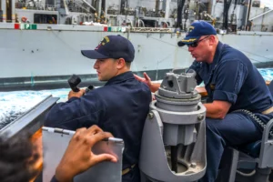 Two sailors in navy uniforms operate equipment and communicate on a ships deck, with a large vessel and churning ocean water visible in the background.