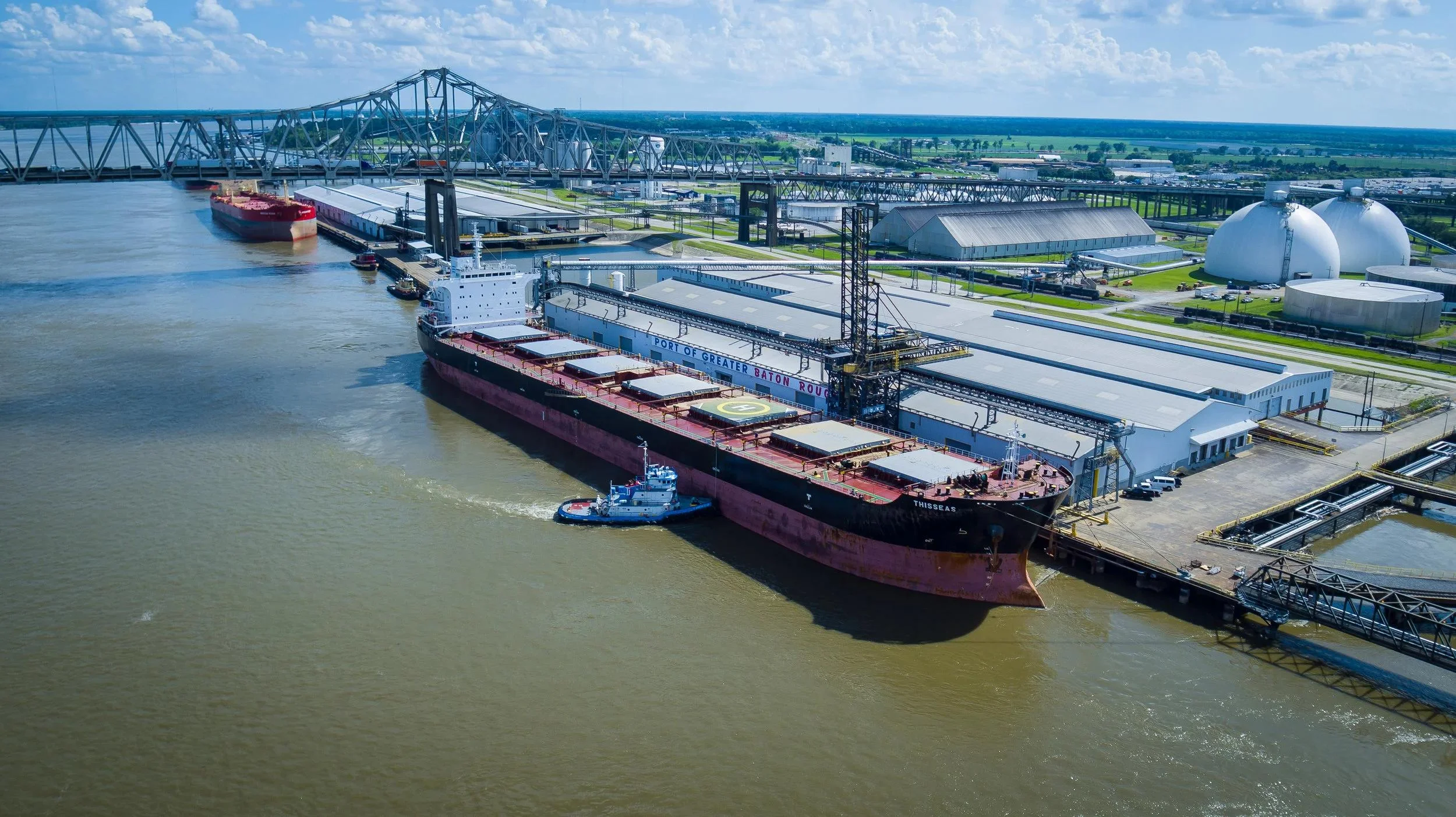 A large cargo ship docked at an industrial port with a tugboat beside it, warehouses, storage tanks, and a bridge in the background under a partly cloudy sky.