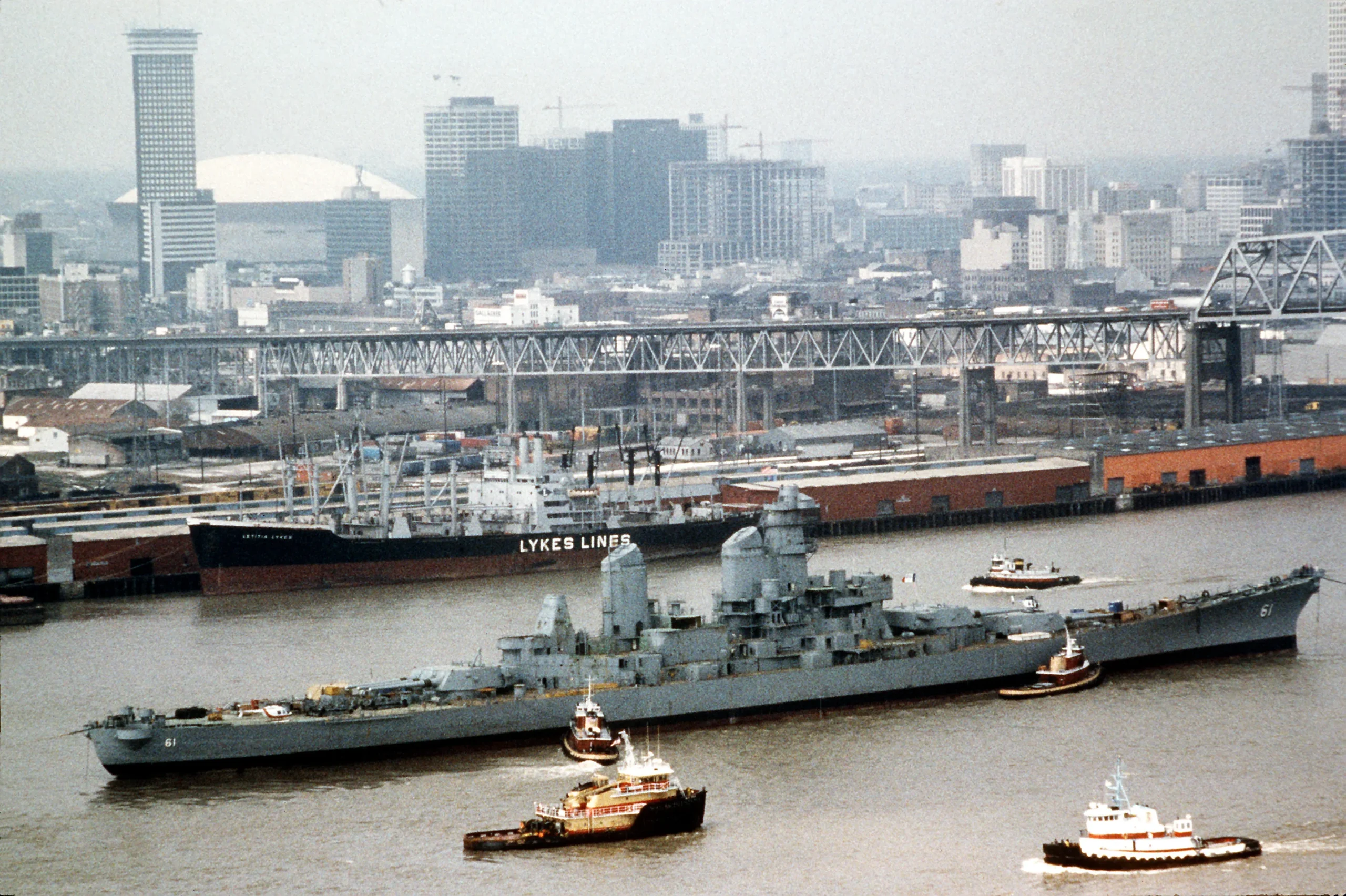 A large gray battleship, identified as the Battleship IOWA, is escorted by tugboats as it moves along a river, with an industrial city skyline and several tall buildings in the background.