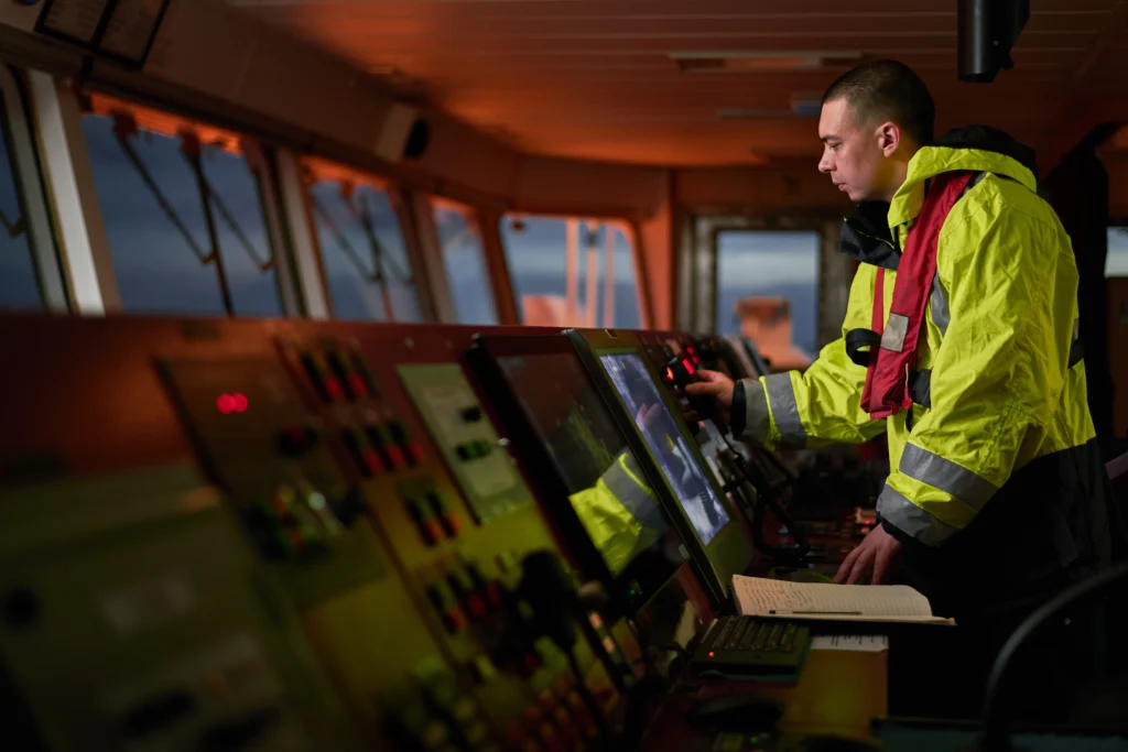 A person in a yellow jacket operates controls on a ships bridge, surrounded by electronic panels and screens, with windows showing an evening or early morning sky outside. A person in a yellow jacket operates controls on a ships bridge, surrounded by electronic panels and screens, with windows showing an evening or early morning sky outside.