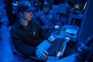 Two uniformed navy personnel work at computer stations in a dimly-lit, blue-lit control room. One types on a keyboard while the other, holding a cup, sits behind and observes. Equipment surrounds them.