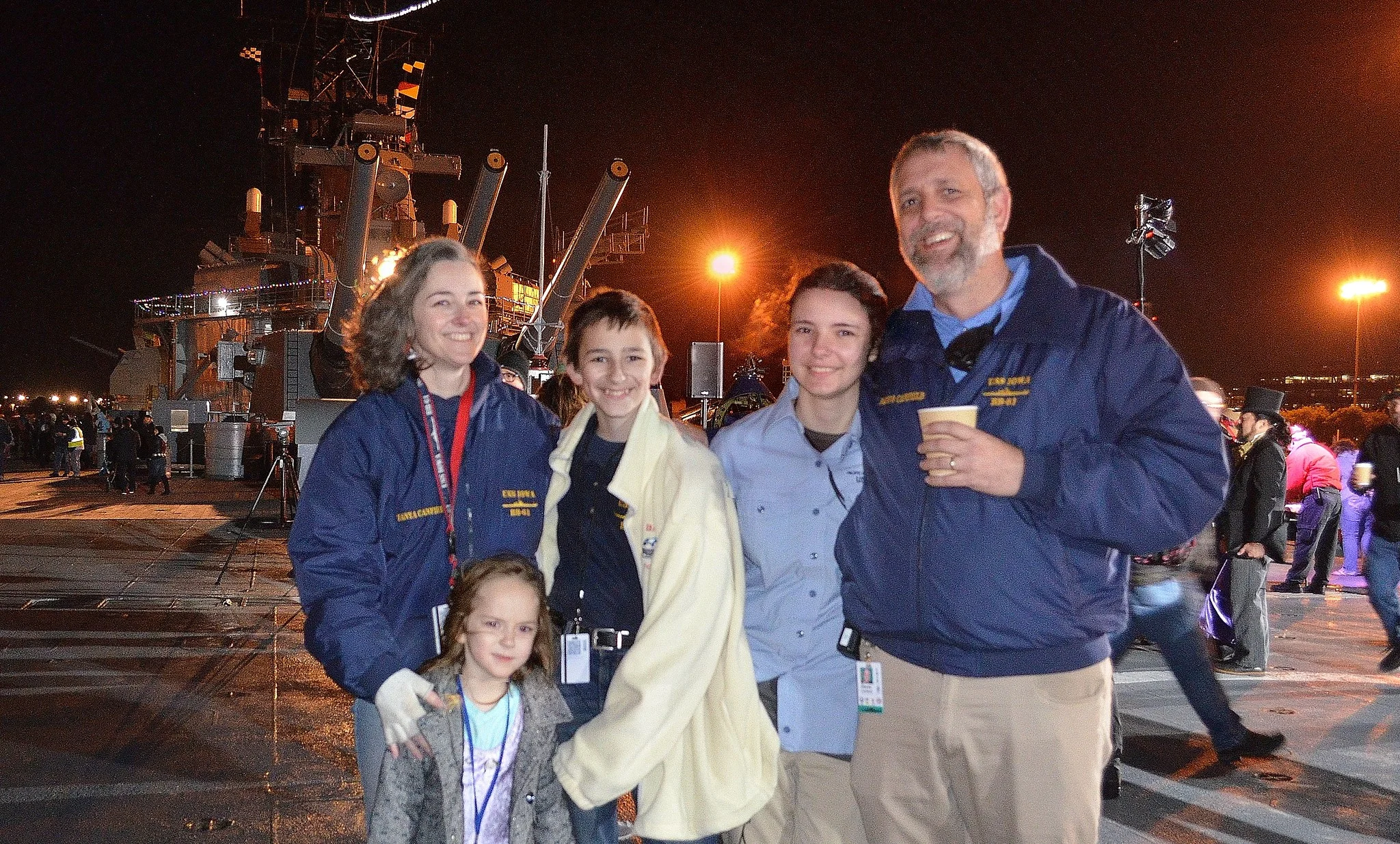 A family of five smiles at night on a ship deck, with the ships equipment visible behind them. Four people wear blue jackets; one child wears a white jacket. Bright lights and other people are in the background.