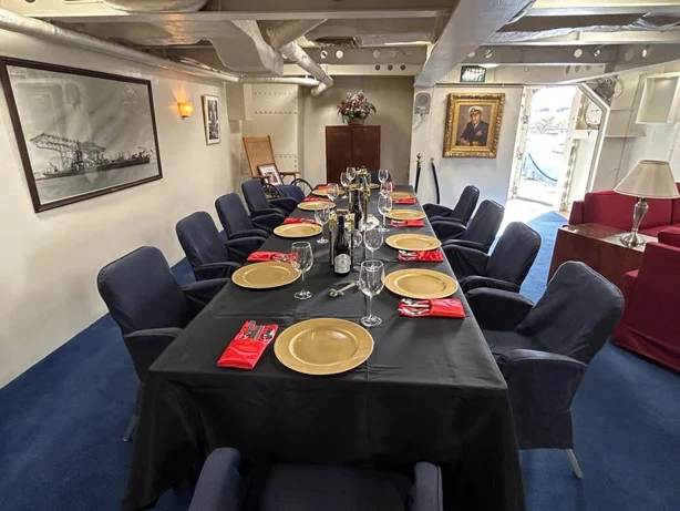 A formal dining table with gold plates, red napkins, and wine glasses is set for twelve in a room with navy blue carpet, cushioned chairs, framed pictures, and a portrait on the wall.