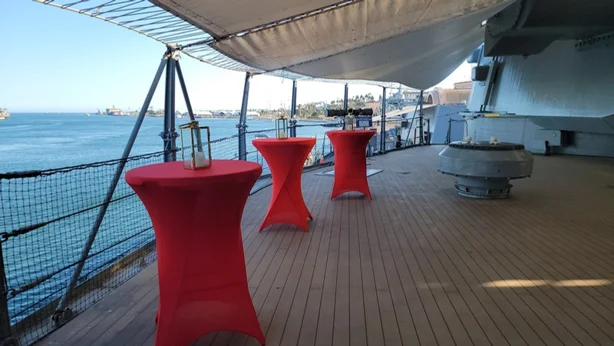 Three tall tables with red covers and lanterns stand on the deck of a ship overlooking the ocean, with blue skies and distant land visible in the background.