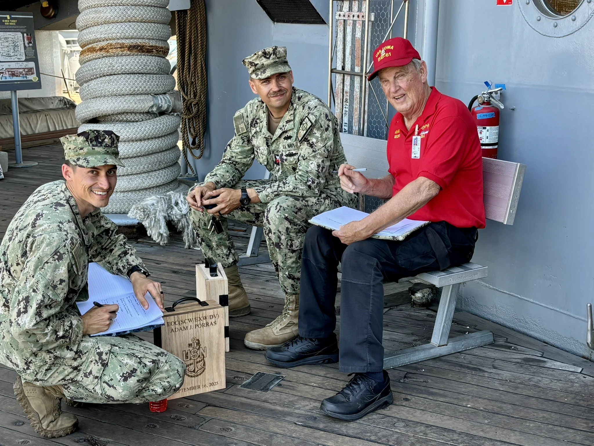 Three men, two in military camouflage uniforms and one in a red shirt and cap, sit on benches on a wooden deck. They are smiling, writing on clipboards, and appear to be in conversation.