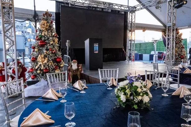 A decorated event space with Christmas trees, a stage with a podium, round tables set with blue tablecloths, napkins, glassware, and a floral centerpiece, under a tented canopy.
