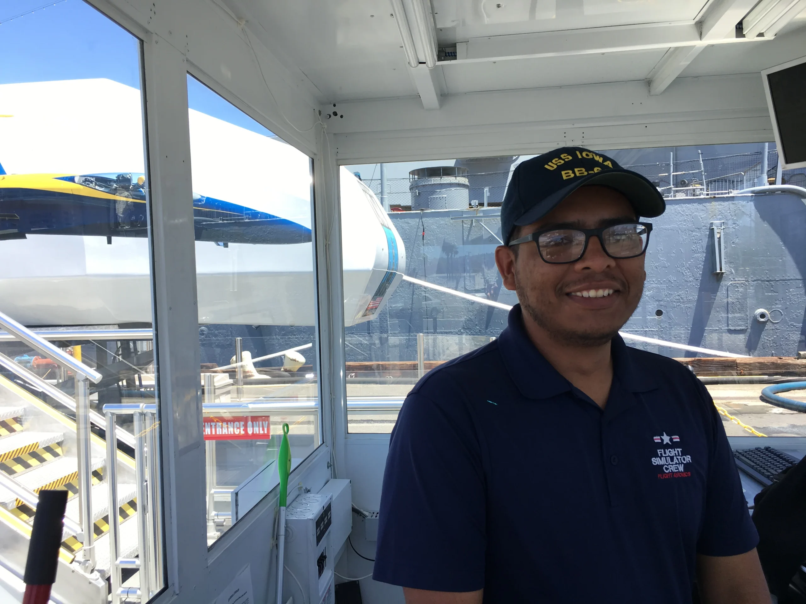 A smiling man in a navy blue shirt and USS IOWA BB-61 cap stands indoors near a window, with part of a jet and a battleship visible outside.