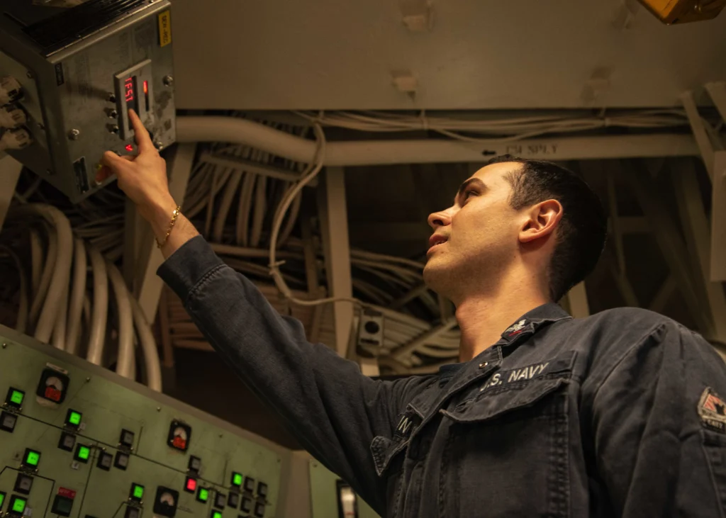 A person in a U.S. Navy uniform operates controls in an industrial setting, reaching up to press a button on a panel surrounded by wires and equipment.