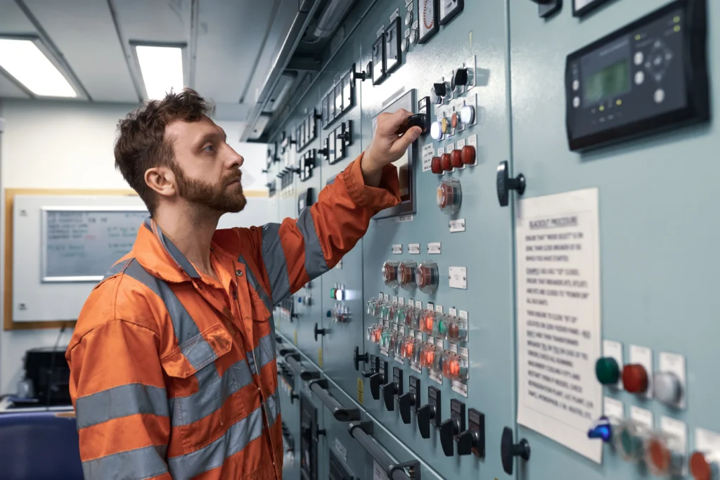 A man in an orange safety suit operates control switches on an industrial control panel filled with buttons, dials, and warning signs inside a brightly lit facility. A man in an orange safety suit operates control switches on an industrial control panel filled with buttons, dials, and warning signs inside a brightly lit facility.