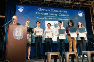 Six students stand on stage holding certificates next to two adults at a podium during the Gravely Experience Student Essay Contest award ceremony. A large blue banner is displayed in the background.