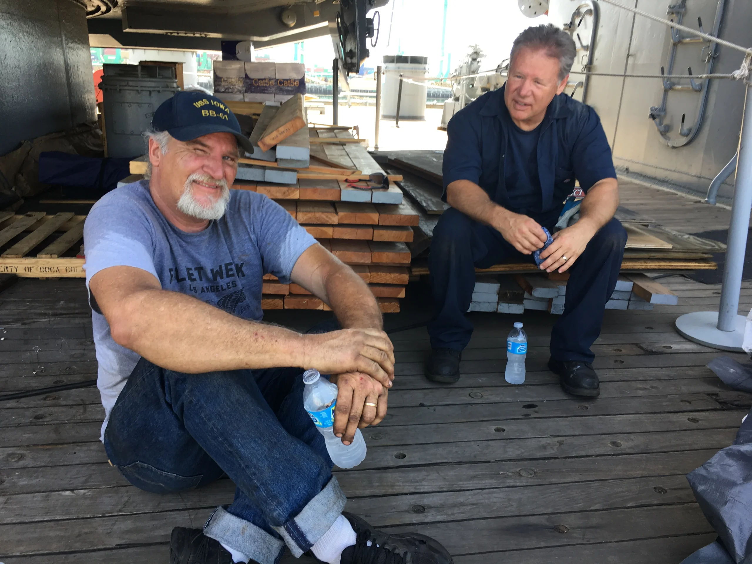 Two men sit on a wooden deck, smiling and holding water bottles. One wears a cap and jeans, the other wears a navy shirt. Stacks of wooden planks and industrial equipment are in the background.