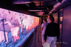 People walk down a corridor beside a long, brightly lit mural depicting ships and a harbor, under exposed pipes and ceiling lights. People walk down a corridor beside a long, brightly lit mural depicting ships and a harbor, under exposed pipes and ceiling lights.