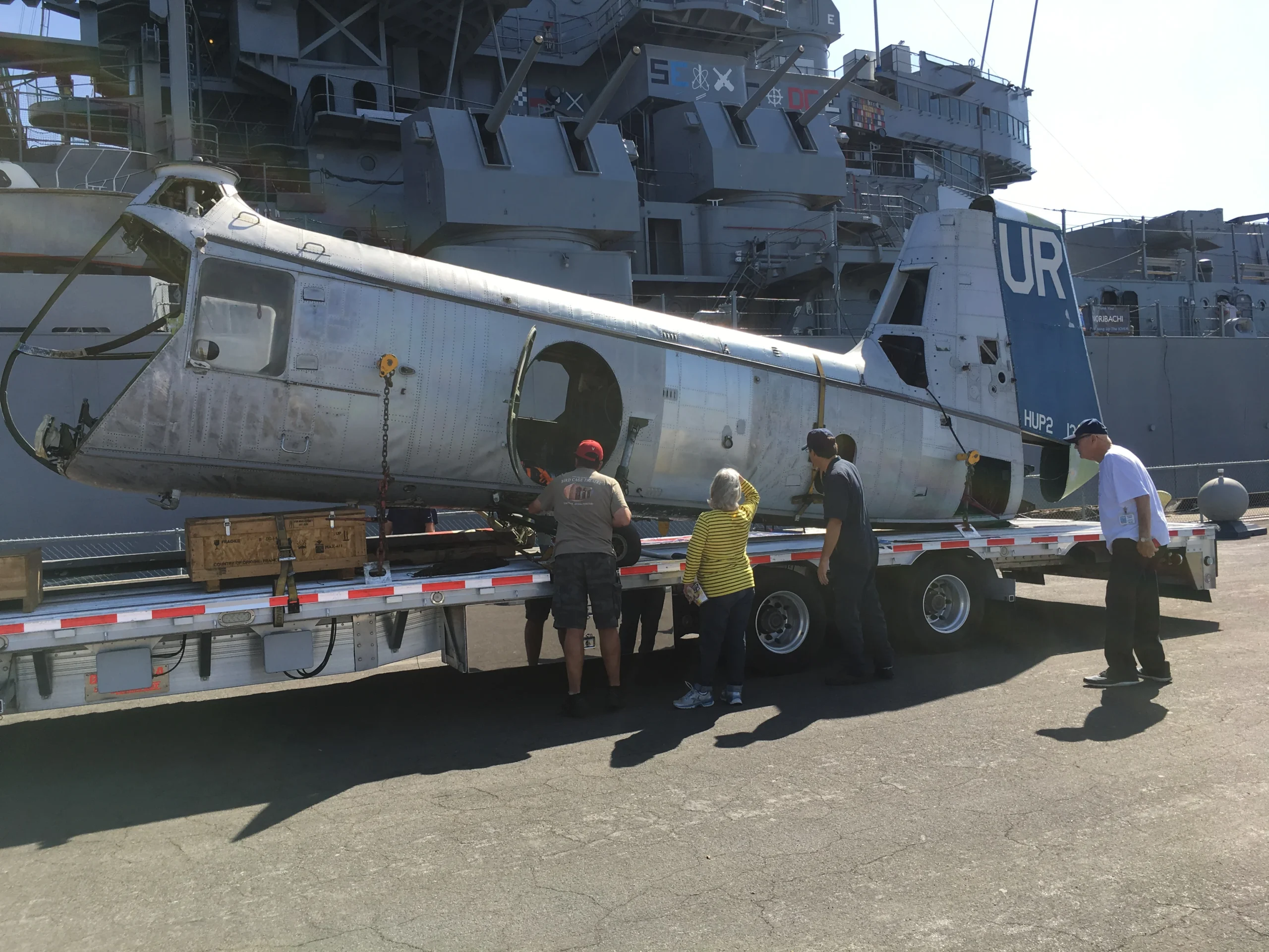 Four people stand by a flatbed trailer carrying the fuselage of a disassembled vintage helicopter. A large Navy ship is visible in the background under bright sunlight.