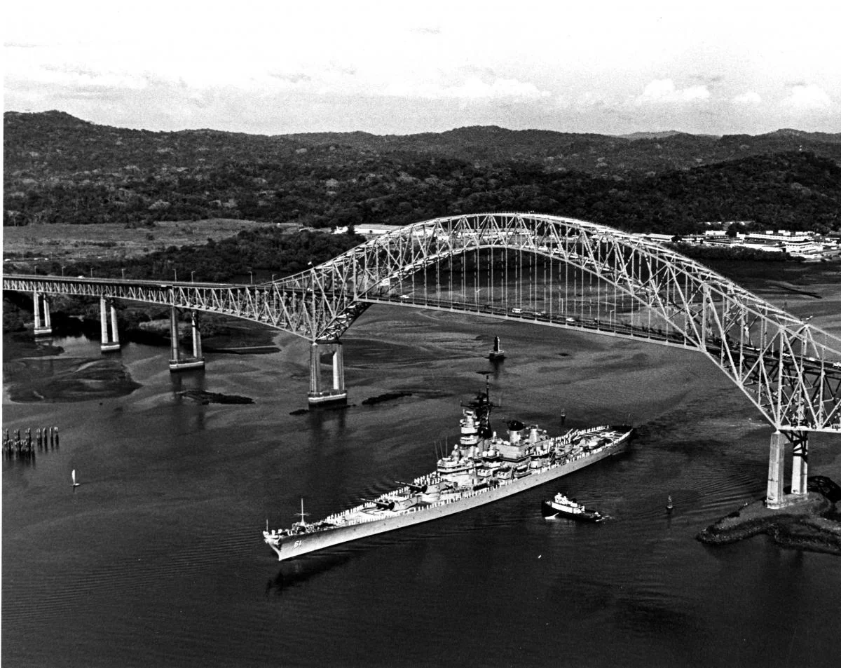 A large naval ship passes under a tall, arched bridge spanning a wide river, with forested hills and scattered buildings in the background.
