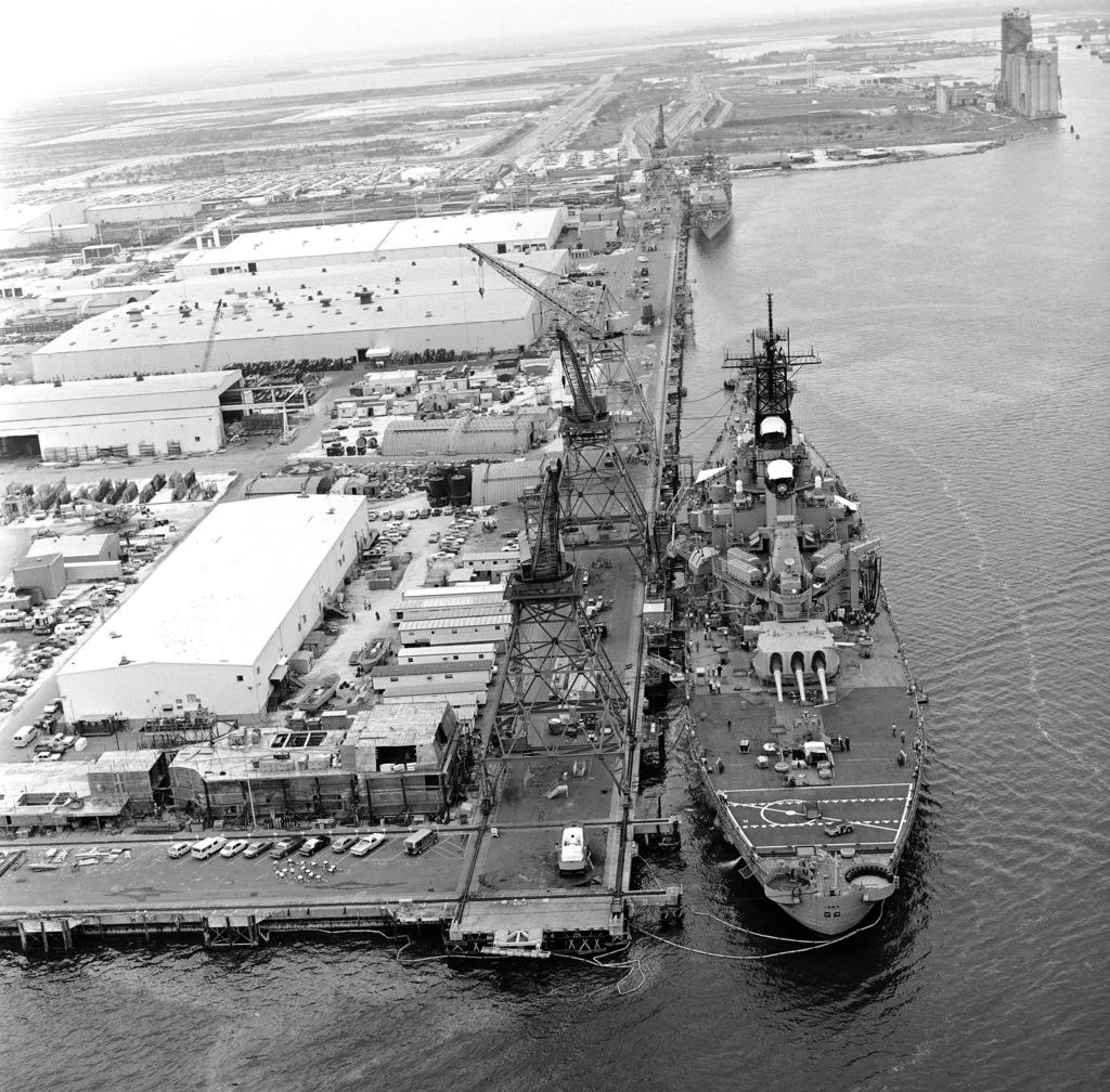 A large battleship docked at an industrial shipyard, surrounded by cranes and warehouses, with cars parked nearby and water on one side. A distant tower is visible across the harbor. Black and white aerial view.