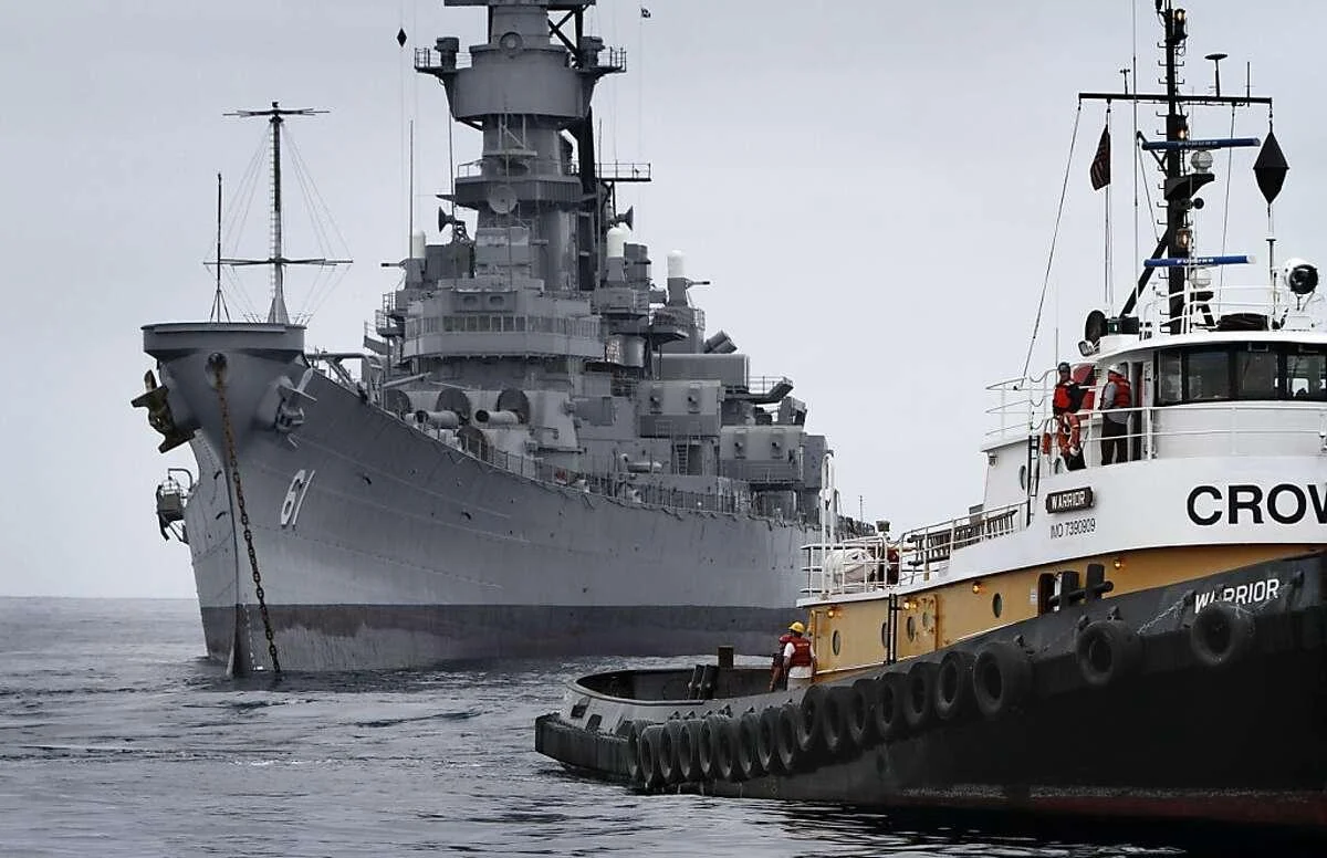 A large gray battleship is anchored at sea, while a smaller black and white tugboat labeled CROW assists nearby under a cloudy sky.