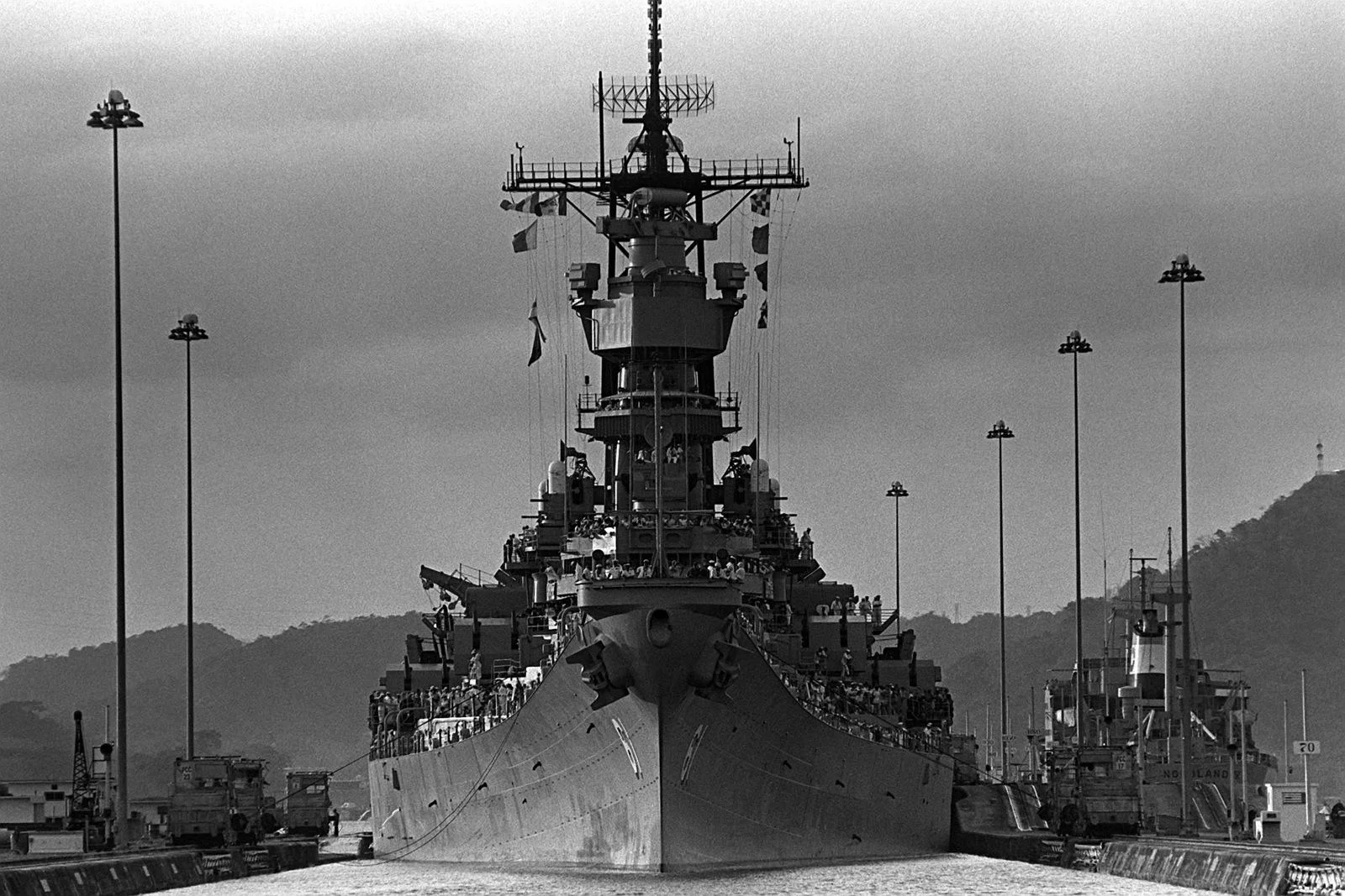 A large battleship passes through the Panama Canal under cloudy skies, flanked by tall light poles and surrounded by misty hills and various industrial structures.