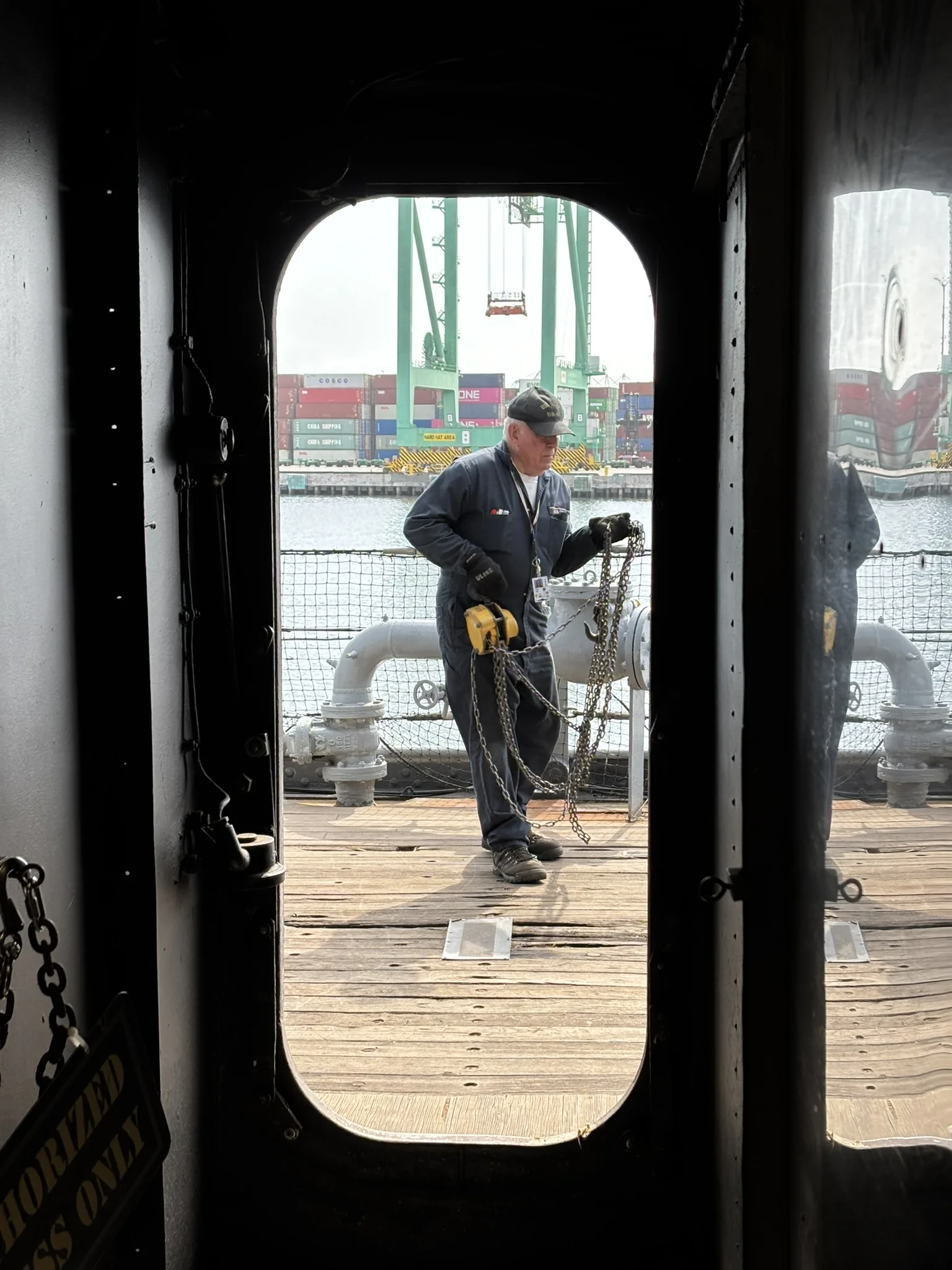 A worker in dark coveralls and a cap handles chains on a docked ship, framed by a doorway. Shipping containers and cranes are visible in the background across the water.