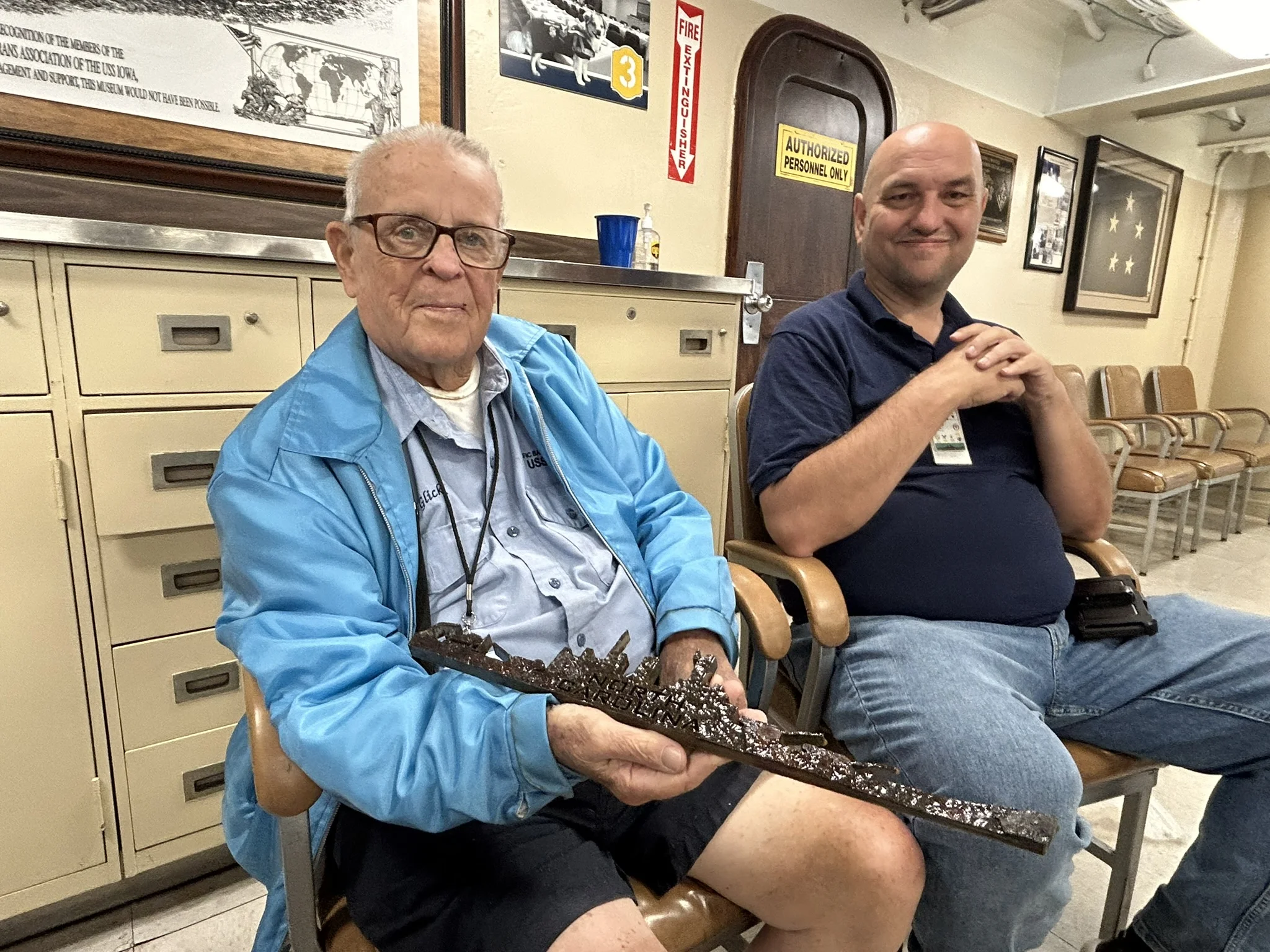 Two men sit in chairs indoors; the man on the left holds a detailed ship model. Both are smiling, and cabinets and framed photos are visible in the background.