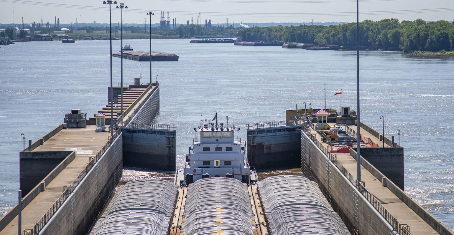 A cargo barge enters a lock on a wide river, with industrial buildings and trees visible in the distant background under a clear sky.