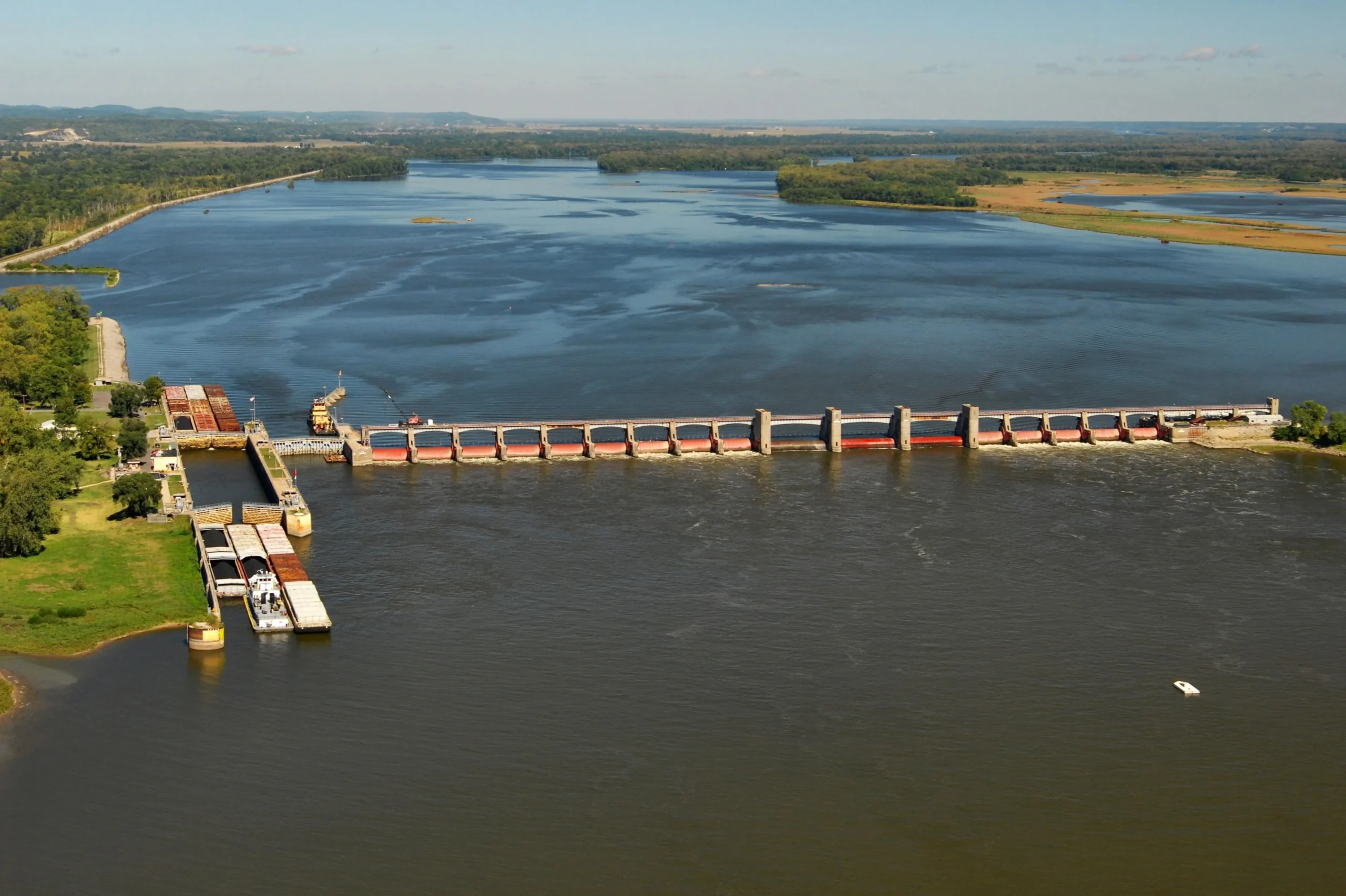 Aerial view of a river with a long dam and lock system; trees and green land border the water, and a small boat is visible in the lower right.
