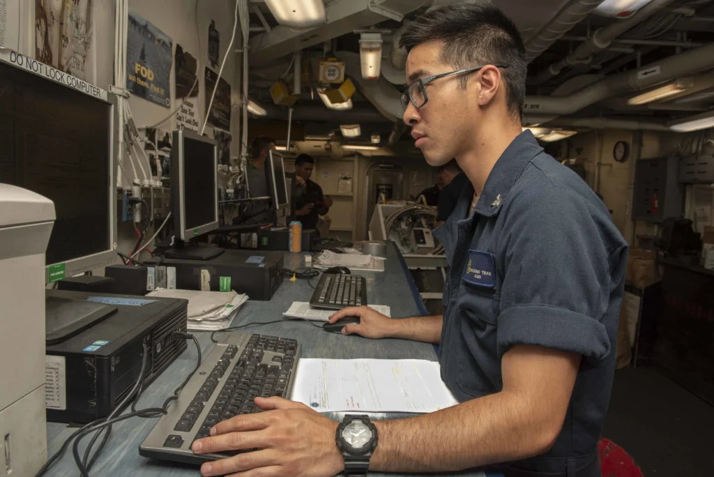 A person in a navy uniform works at a computer desk in an office with multiple monitors, keyboards, and documents, inside what appears to be a ship or industrial setting.