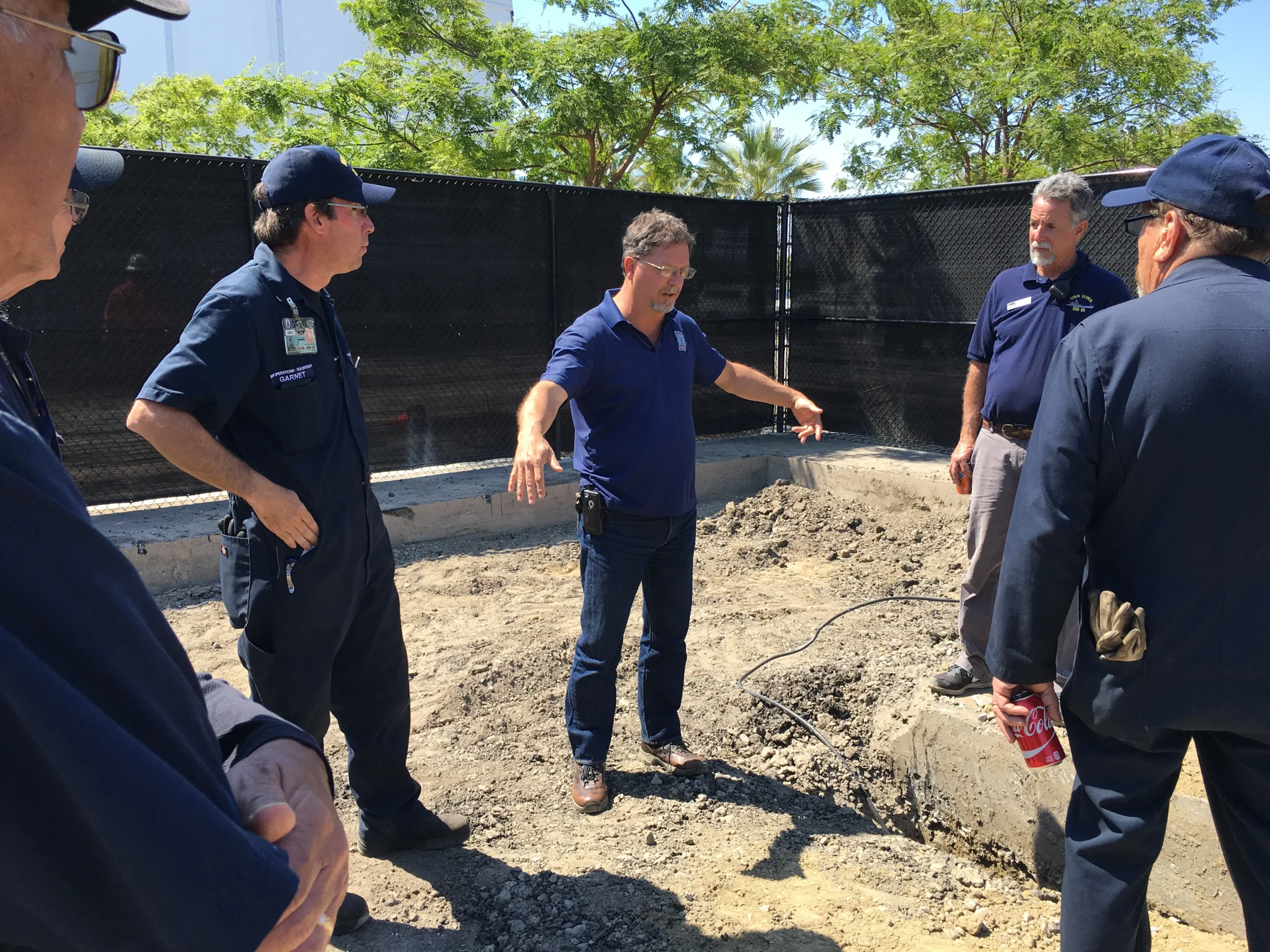 A group of men, some in uniforms, stand and talk at a construction site with dirt ground and fencing. One man in a blue shirt gestures with his arms while others listen. Trees and sunlight are visible in the background.