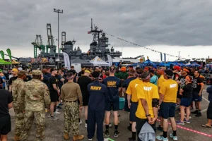 A large group of people, many wearing NAVY shirts, gathers outdoors near a battleship and tents under a cloudy sky. Some individuals are in military uniforms.