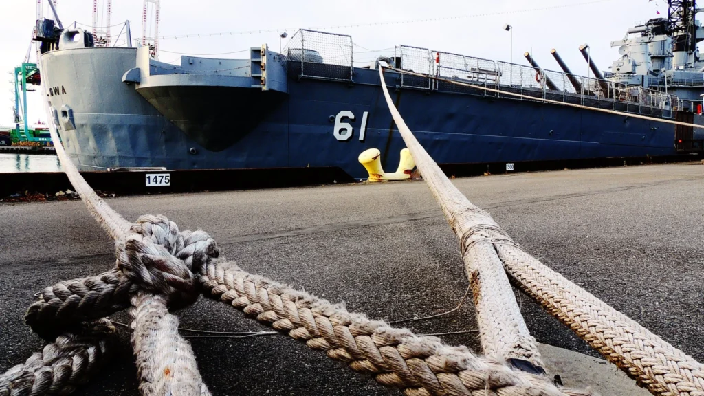 Close-up of thick mooring ropes tied to a dock, securing a large gray battleship with the number 61 on its hull. The ship has visible turrets and is docked at a harbor.