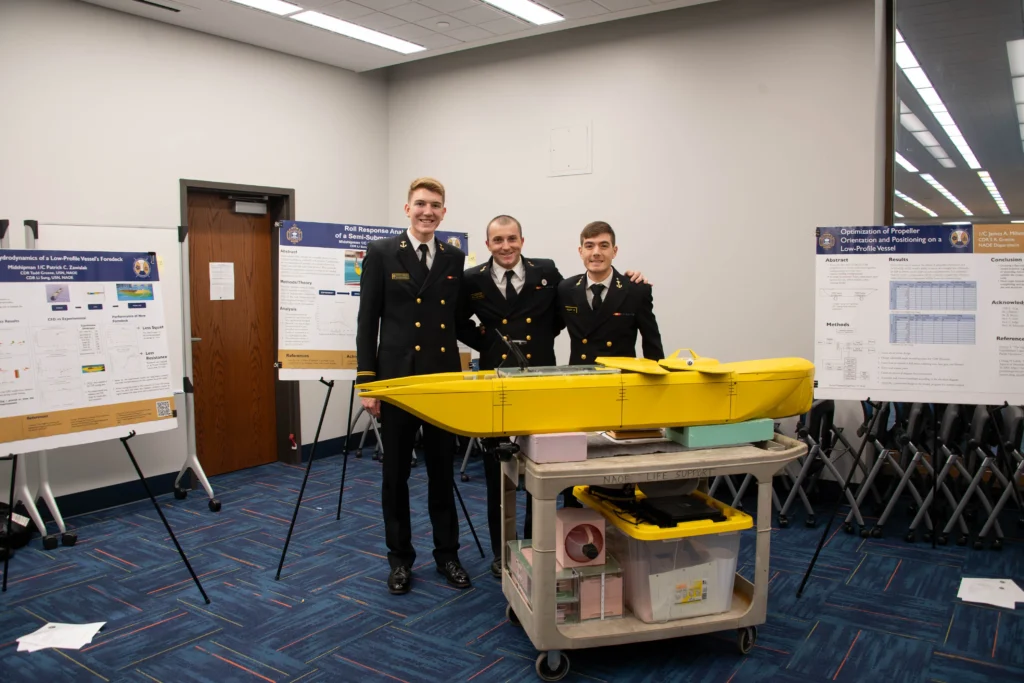 Three people in naval uniforms stand smiling behind a yellow model boat on a cart, with scientific posters displayed on easels in the background. The scene appears to be at a research or academic presentation event focused on Ship Design, offering insights into Naval Architecture careers.