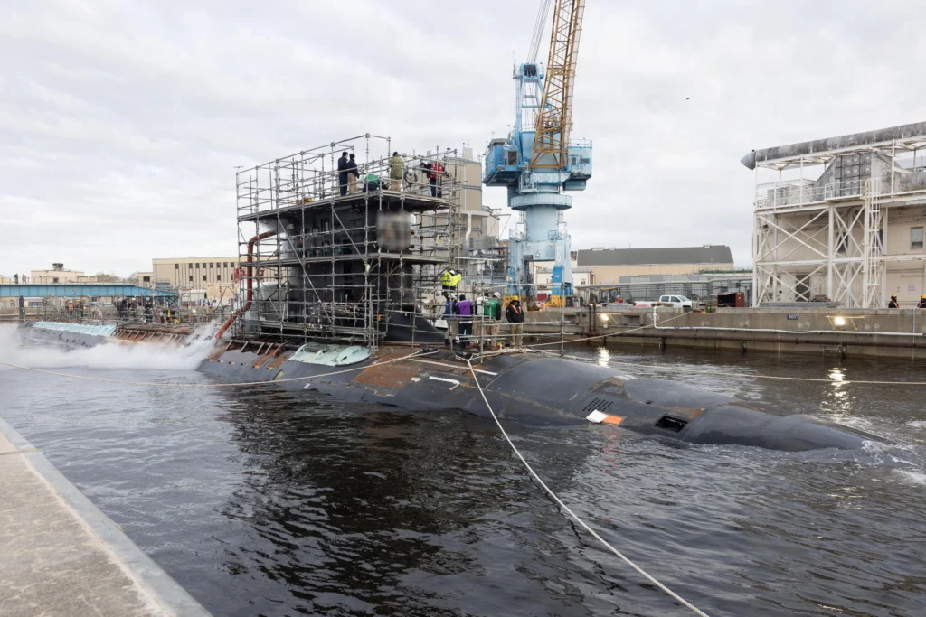 A submarine docked at a shipyard is surrounded by scaffolding and workers wearing safety gear, with cranes and industrial buildings visible in the background. Steam or smoke rises from the area.