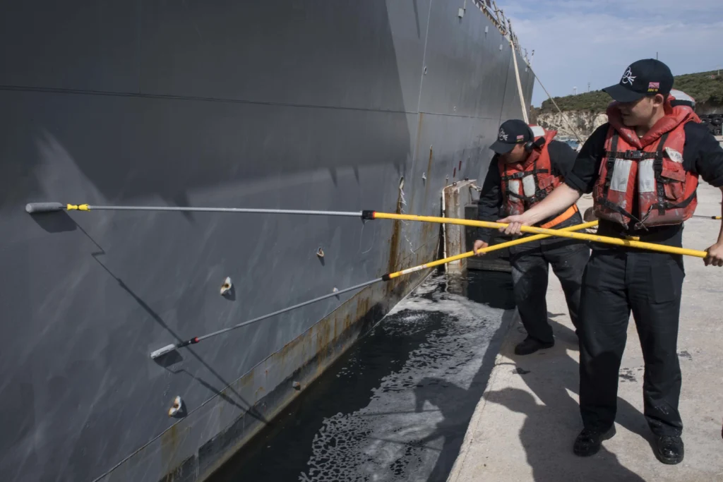 Two people wearing life vests use long poles with brushes to clean the side of a docked ship. The ships hull shows some rust and algae near the waterline. The individuals appear focused on their task.