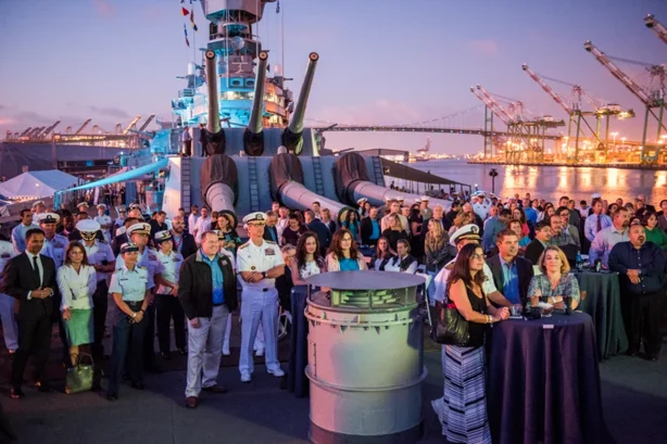 A large group of people, including military personnel in uniform, stand on the deck of a battleship at sunset. Cranes and a harbor are visible in the background.
