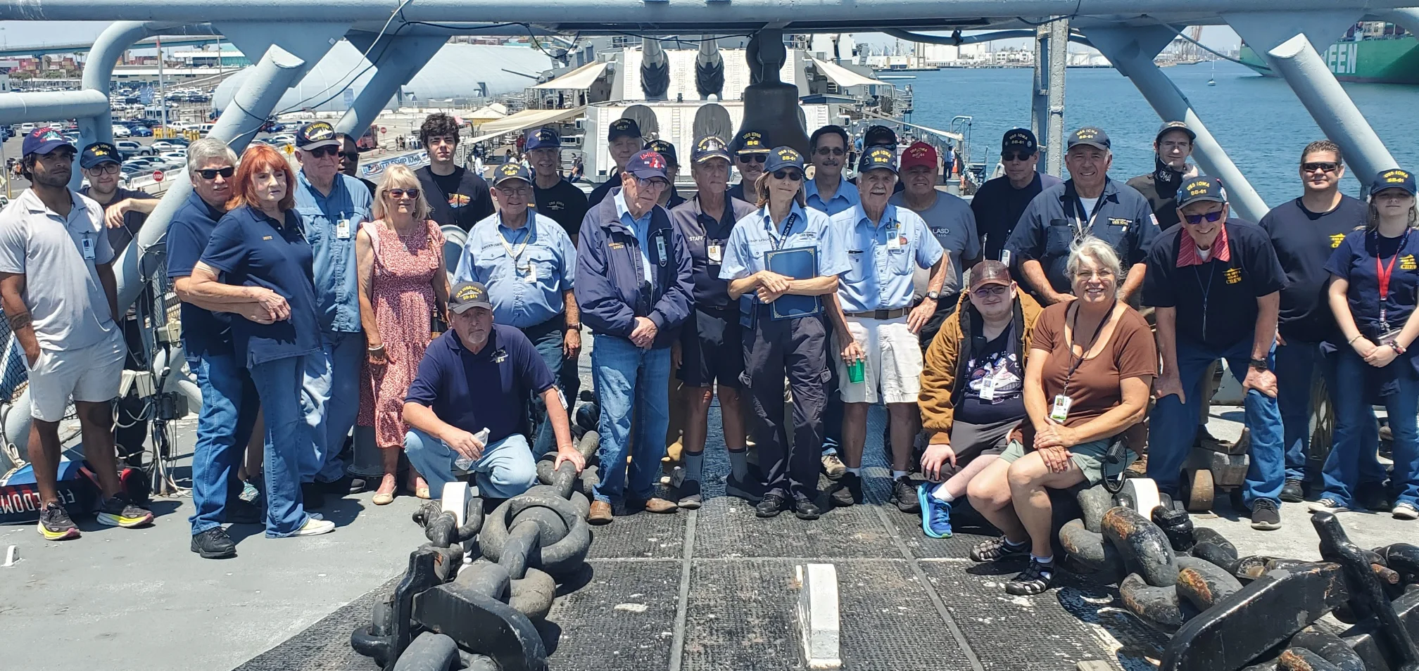 A group of people, some in navy uniforms and others in casual clothes, stand and sit on the deck of a ship at a port on a sunny day. Large chains and a harbor are visible in the background.