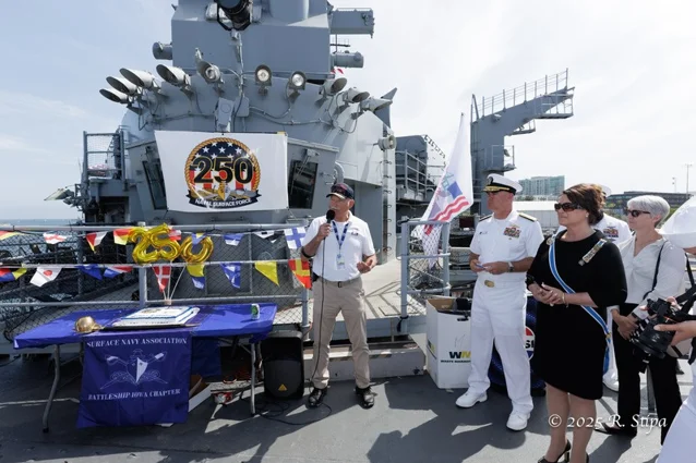 A group of people stands on a battleship deck near a table and 250 anniversary decorations, with a speaker addressing the group and naval officers present. A group of people stands on a battleship deck near a table and 250 anniversary decorations, with a speaker addressing the group and naval officers present.