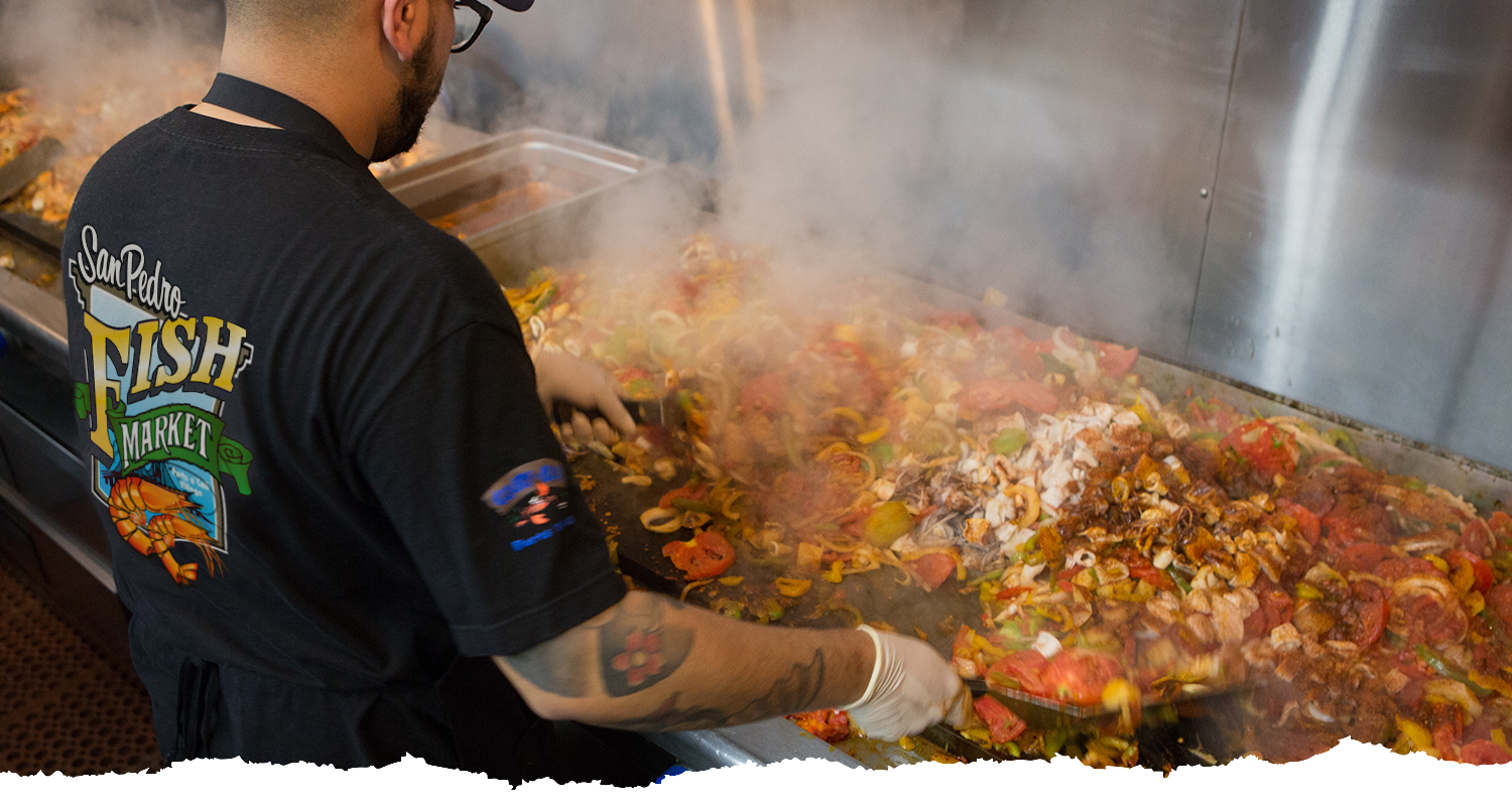 A person in gloves cooks a colorful seafood and vegetable mixture on a large hot grill, with steam rising up. The shirt reads San Pedro Fish Market.. A person in gloves cooks a colorful seafood and vegetable mixture on a large hot grill, with steam rising up. The shirt reads San Pedro Fish Market..
