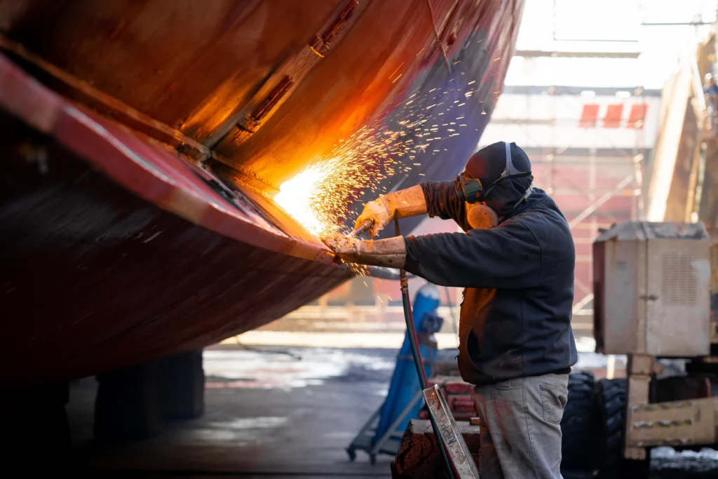 A worker in protective gear uses a welding torch on the side of a large metal structure, emitting bright sparks. The scene takes place outdoors, likely in a shipyard or industrial area.
