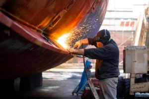A worker in protective gear uses a welding torch on the side of a large metal structure, emitting bright sparks. The scene takes place outdoors, likely in a shipyard or industrial area.