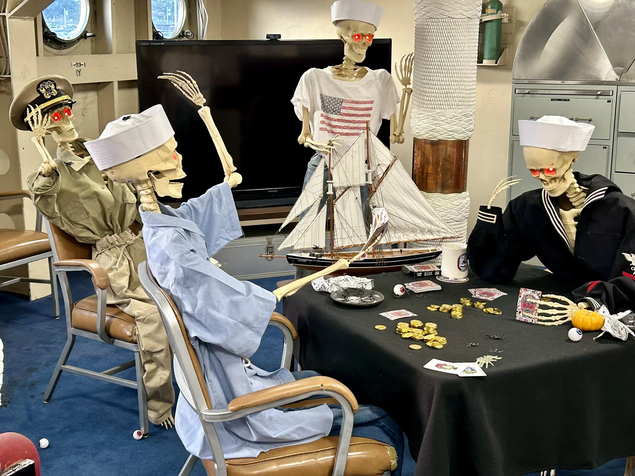 Four skeletons dressed as sailors and a captain sit around a table with cards, coins, and pirate-themed items. One raises its arm, and all have glowing red eyes. A model ship is displayed in the background.