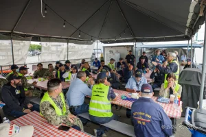A large group of people in safety vests and uniforms sit at tables with checkered cloths under a canopy, listening to a speaker standing near the front. The setting appears to be outdoors.