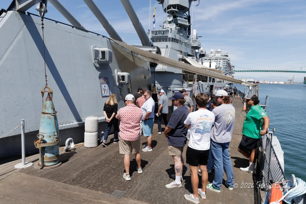 A group of people listens to a guide on the deck of a large gray ship docked by a harbor, with equipment and masts visible under a sunny sky.