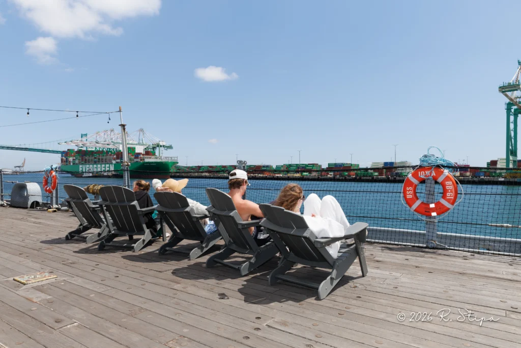 Five people relax in Adirondack chairs on a wooden pier by the water, with cargo ships, cranes, and shipping containers visible in the background under a sunny sky.