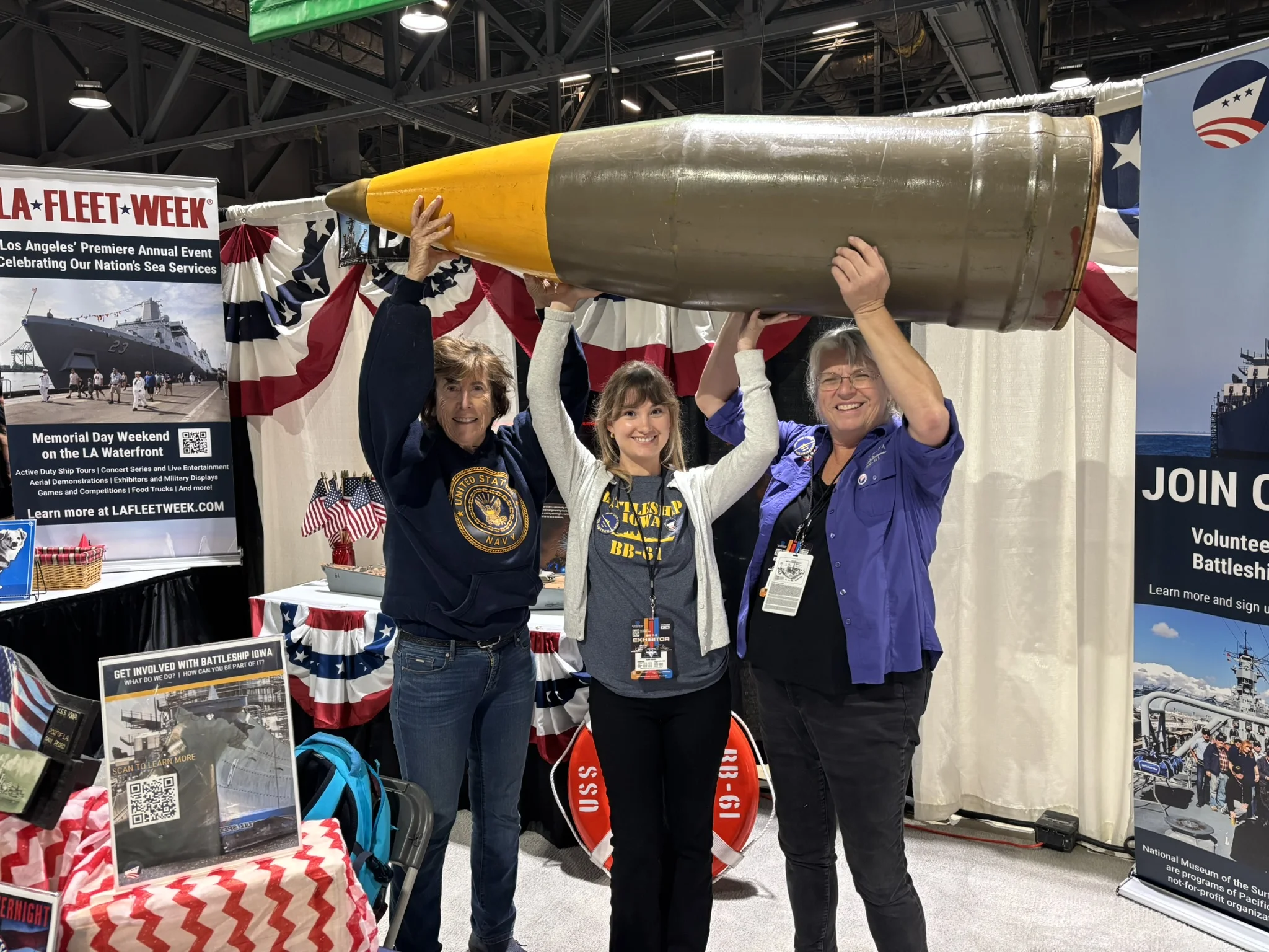 Three smiling women stand indoors at a patriotic-themed booth, holding a large replica artillery shell above their heads, surrounded by banners, brochures, and informational displays about battleships and Fleet Week.