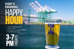 A glass of beer with a Battleship IOWA logo is in focus, with cranes and water blurred in the background. Text promotes Vicky’s Happy Hour at Vicky’s Doghouse—Fridays from 3-7 PM—featuring games, music, sports, and stunning waterfront views.