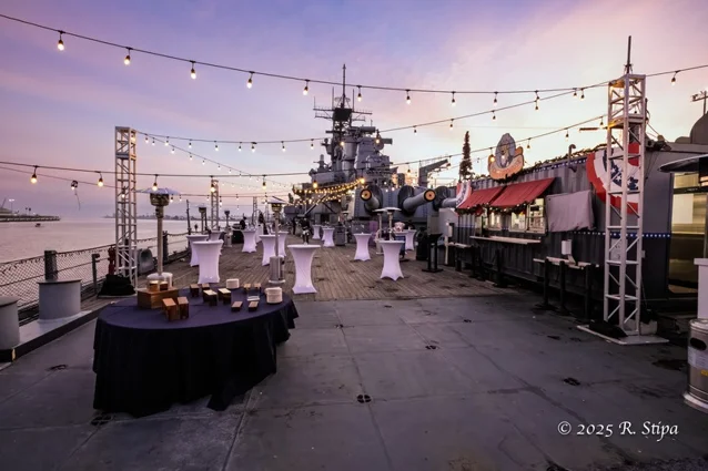 An outdoor event setup with cocktail tables, string lights, and vendor booths on the deck of a battleship at sunset. The ships large guns and harbor are visible in the background.