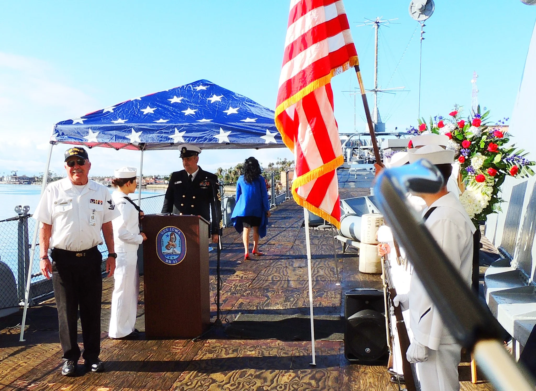 Several people in naval uniforms stand on a ships deck under a star-patterned canopy. An American flag is in the foreground, and a woman in a blue dress walks toward a podium with floral wreaths nearby.